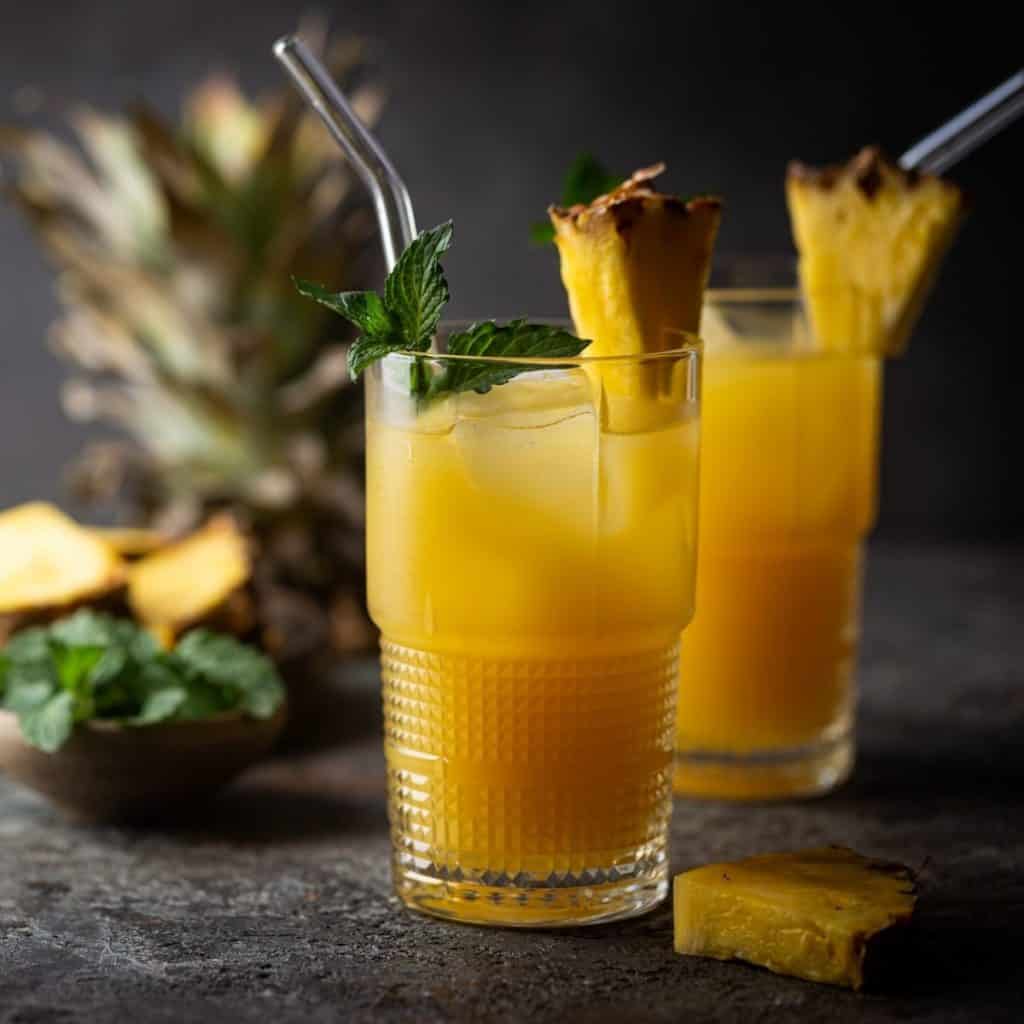 Two bright vodka and pineapple juice cocktails in front of pineapple slices and fresh herbs.