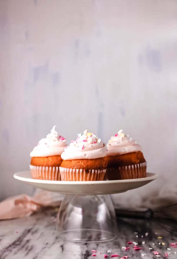 3 pink frosted cupcakes with sprinkles on a cake plate. 