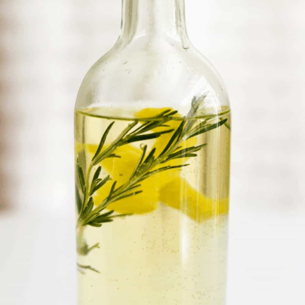 Close up of a bottle of white wine with lemon peel and rosemary sprigs in it.