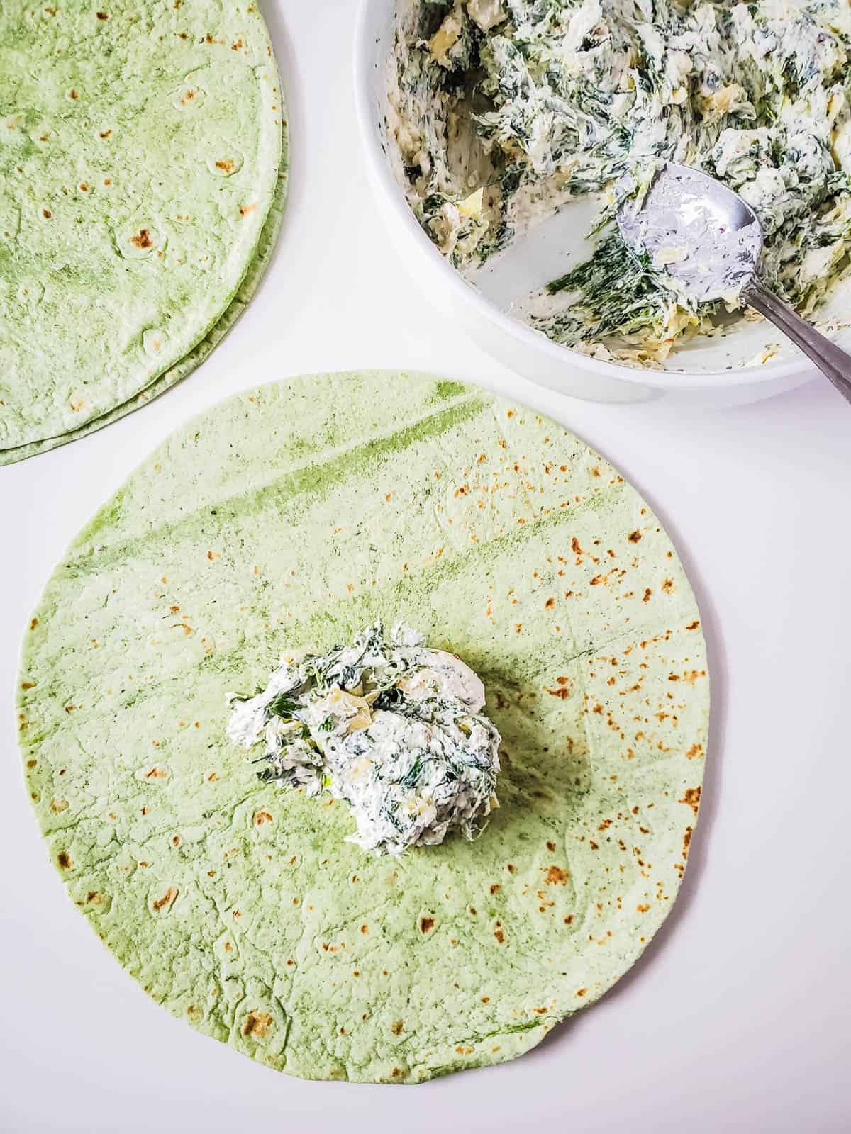 Topping a spinach tortilla with spinach and artichoke dip.