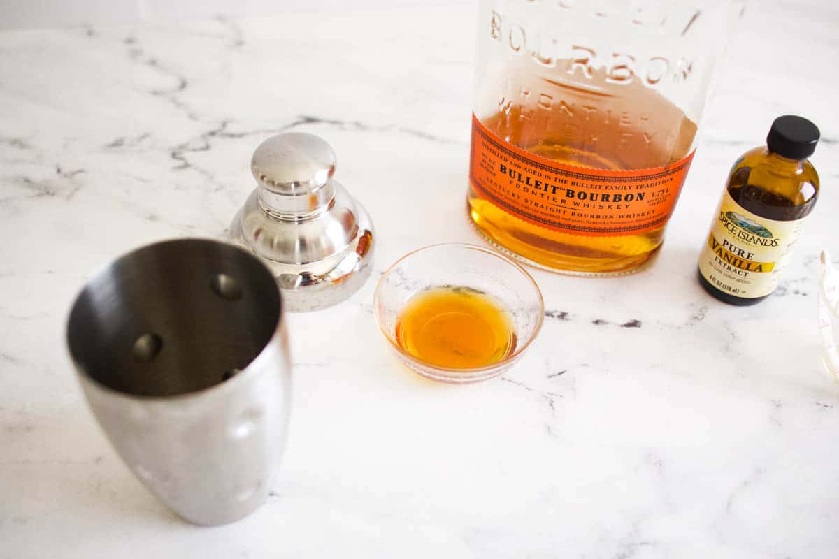 A bottle of bourbon on a table next to a small glass bowl of honey next to a cocktail shaker. 