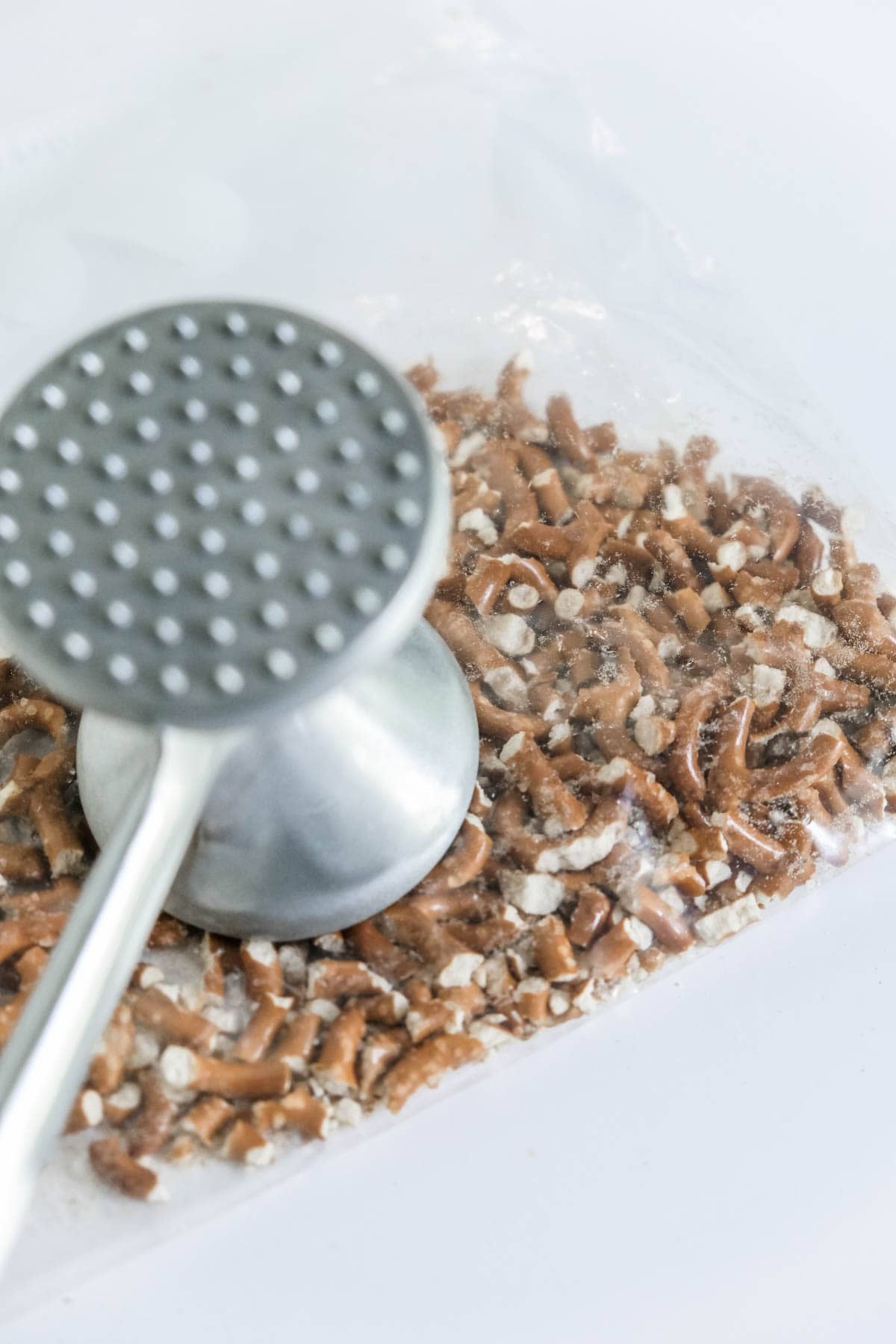 Crushed pretzels in a plastic bag with a meat tenderizer being used to crush them. 