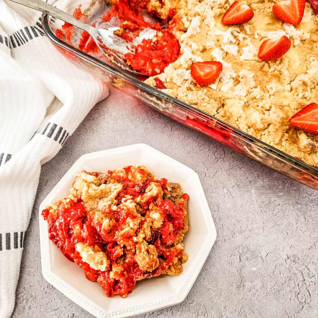 Strawberry Cheesecake Dump Cake in a baking dish with a serving in a white bowl.