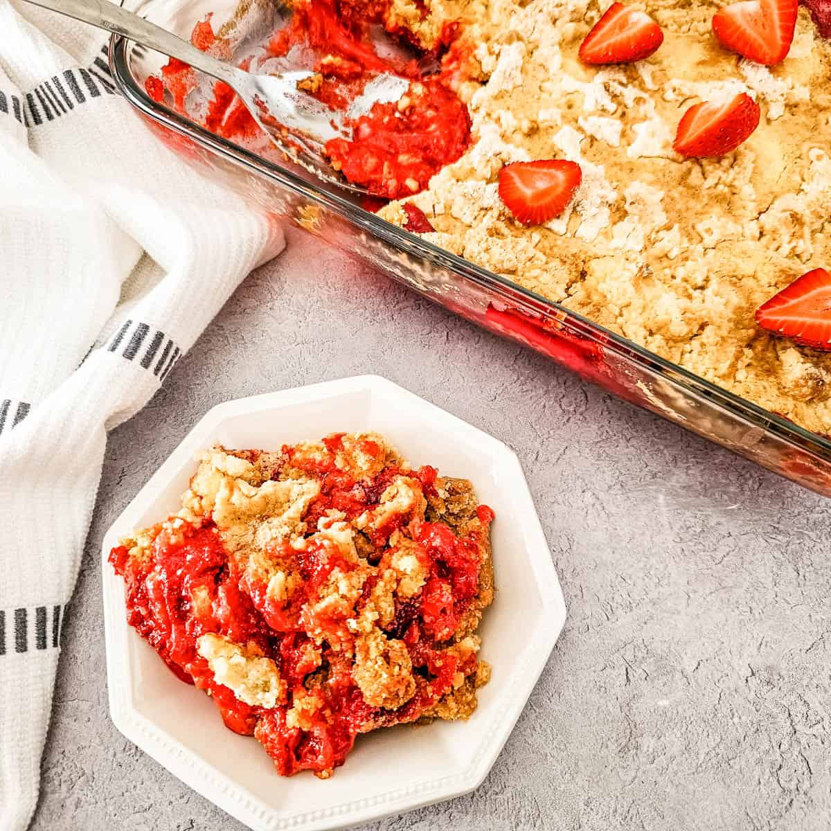 Strawberry Cheesecake Dump Cake in a baking dish with a serving in a white bowl.