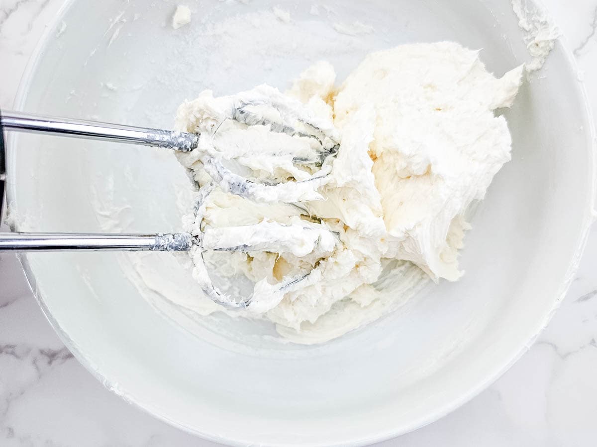 Softened cream cheese and sugar in a mixing bowl with hand mixer attachments.